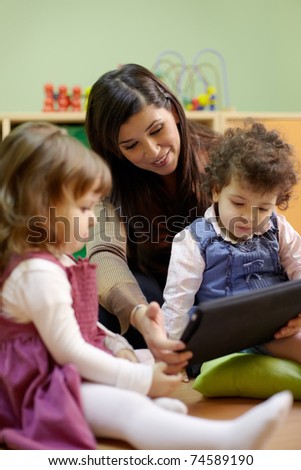 Caucasian female educator and little girls reading fairy tale on touch pad computer in kindergarten