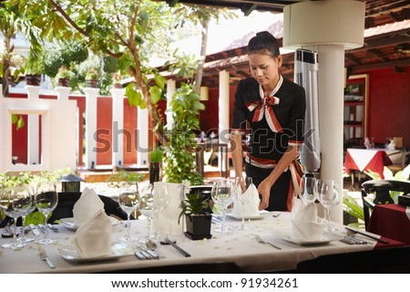 Attractive young woman working as waitress in exclusive restaurant, setting up a table. Waist up, front view