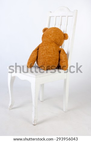 Old brown teddy bear sits on a chair, his back to us