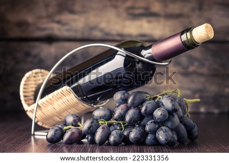 Winery background. Bottle of red wine in holder with cluster of dark blue grapes on wooden table. Toned image