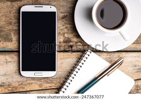 Smartphone with notebook and cup of strong coffee on wooden background. Cell phone with writing set with espresso