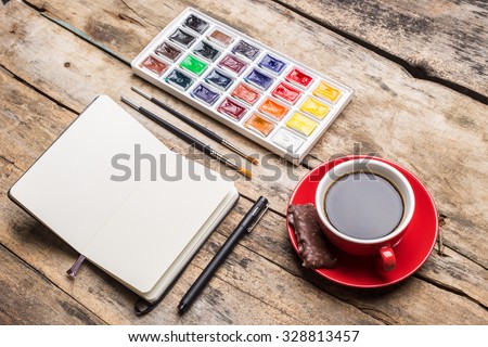 Opened notebook with watercolour paints and cup of coffee on wooden table. Angle view artistic mock up
