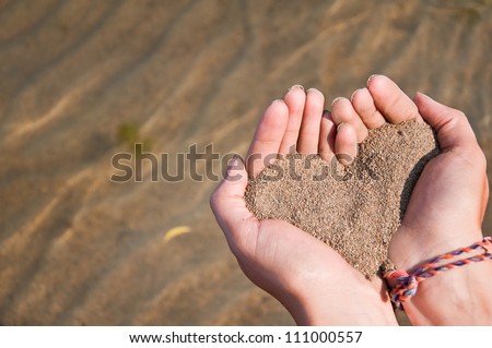 Heart made of sand in a girls hands above crystal clear water at the beach