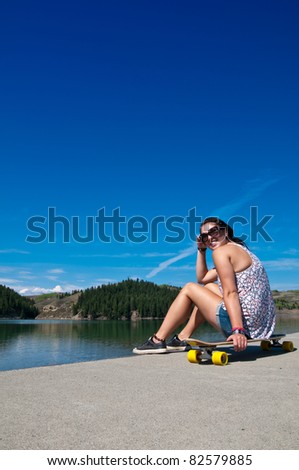 Young adult girl sitting on her long board by a lake looking back at the camera & smiling on a sunny summer day
