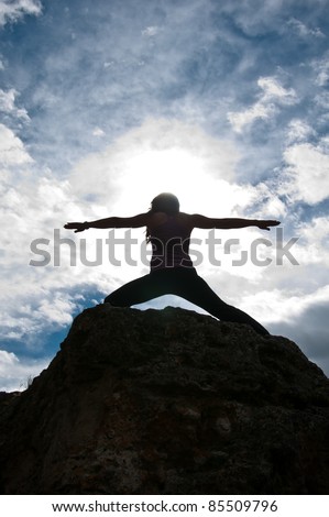 Silhouette of a young attractive girl doing the Warriors pose from yoga on top of a rock with the sky and clouds behind her.