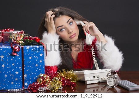 Stressed beautiful Santa girl sitting at desk and speaking on phone surrounded by Christmas presents, decorations, alarm clock and calculator over dark background.