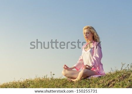 Beautiful blonde young lady wearing pink clothes meditating in yoga pose early in the morning in nature.