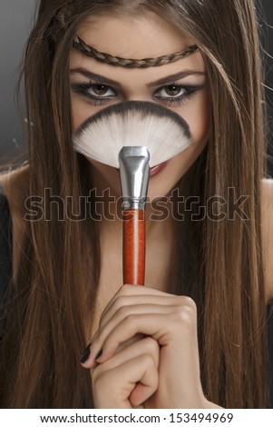 Young happy lady holding a big makeup brush with both hands in front of her face while looking at the camera. Professional make-up.