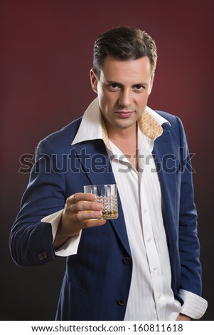 Portrait of serious elegant young man in suit toasting with glass of alcohol against dark red background.