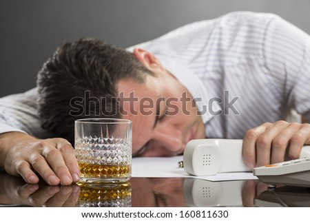 Portrait of tired overwhelmed young man sleeping at work with glass of alcohol on desk. Selective focus on the glass.