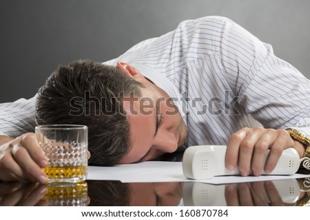 Portrait of tired overwhelmed young man sleeping at work with glass of alcohol on desk.
