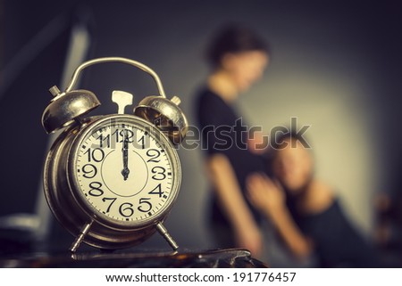 Pregnant expecting woman being caressed on her tummy by her lady friend with alarm clock in the foreground. Selective focus on the alarm clock.