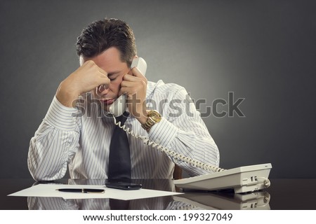 Overwhelmed businessman in white shirt and tie having a headache during a stressful phone conversation. Tired thoughtful businessman with one hand on his forehead taking a tedious phone call.