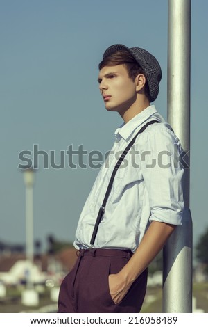 Side view of a thoughtful young guy posing with hand in pocket and rolled up sleeves, wearing hat, shirt and trousers with suspenders, looking away while leaning his back against a street lamp pillar.