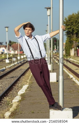 Full body of a pensive anxious young man wearing hat, shirt with rolled up sleeves and trousers with suspenders, looking into the distance while holding on a street lamp post on the railway platform.