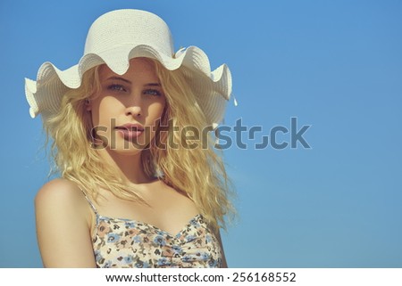 Portrait of beautiful young lady with long blond curly hair and blue eyes wearing white sun hat with wavy brim over clear blue sky. Copy space.
