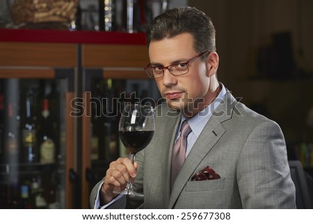 Portrait of sophisticated confident wealthy gentleman in stylish suit toasting with glass of wine against blurred restaurant background.