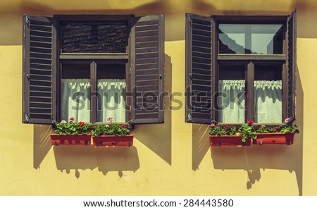 Window with wide open brown wooden shutters and decorative blooming flowers in pots, against bright yellow wall.