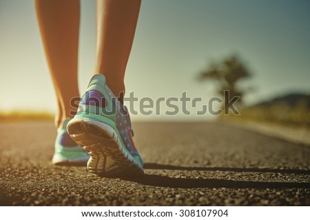 Closeup of female runner shaved feet in running shoes going for a run on the road at sunrise or sunset. Shallow depth of field, toned with instagram like filter, flare effect.