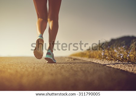 Woman feet in running shoes going for a run on the road at sunrise or sunset. Shallow depth of field, low angle, toned with warm instagram like filter, flare effect.