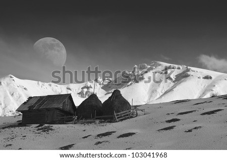 Big moon and old cottage and hay stacks in snow covered mountains, black and white composition