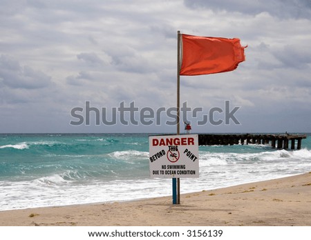 Color DSLR landscape picture of red warning flag and danger sign at the beach with rough surf, a dramatic sky background and approaching wind storm. Horizontal orientation with copy space for text
