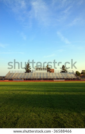 Color DSLR image of Americanl sports football stands or bleachers, as seen from green grass field at dusk with shadows and sunlight; clear, blue sky background. Vertical with copy space for text.