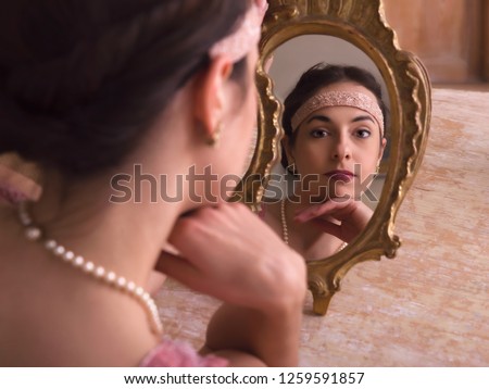 Sensual young woman in 1920s flapper dress and headband looking in an antique mirror