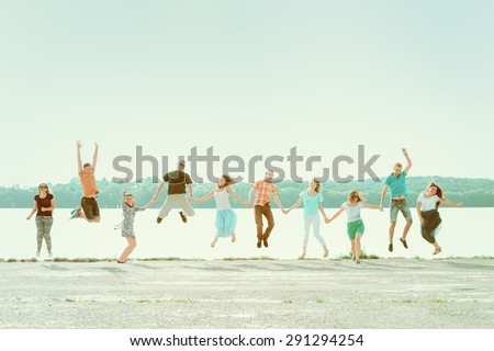 Group of people holding hands and jumping at the park near the lake. Smiling girls and boys having fun