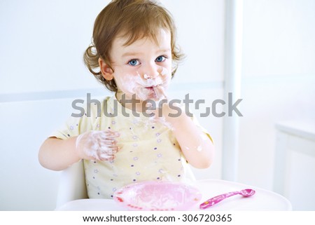 Adorable baby girl playing with food. Child eating yogurt. Dirty face of happy kid. Portrait of a baby eating with a stained face.