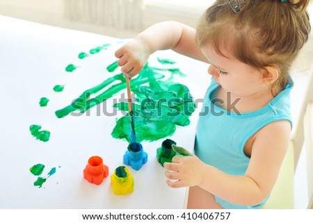 Cute little toddler child painting with paintbrush and colorful paints. Adorable baby girl drawing on white paper near window in light room