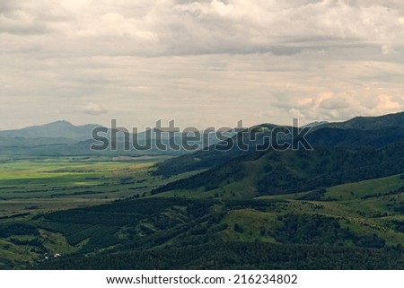 Aerial view of mountains, hills and flat land with forest, agricultural fields, farms and villages in the Altai region (Russia) by a dull cloudy summer day.