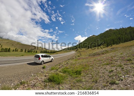 Rural mountains landscape with hills, mountains, road, blue summer sky with clouds and sun and car parked at the roadside during a trip over Altai region (Russia).