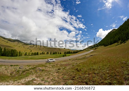 Rural mountains landscape with hills, mountains, road, blue summer sky with clouds and sun and car parked at the roadside during a trip over Altai region (Russia).