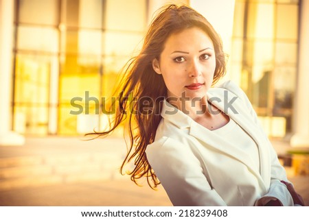 Portrait of attractive young businesswoman with long dark hair and red lips in front of building at sunset.