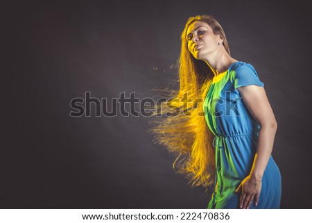 Beautiful female model in blue dress posing at studio in the light flashes on black background. Attractive cute girl with long sun-drenched blowing and  flying flaming hair.
