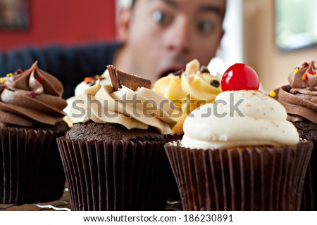 Close up of some delicious gourmet cupcakes frosted with a variety of frosting flavors.  Shallow depth of field with the face of a hungry man lurking in the background.