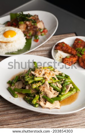 Freshly prepared Asian style chicken and asparagus stir fry with garlic and a variety of other Thai food dishes in the background.