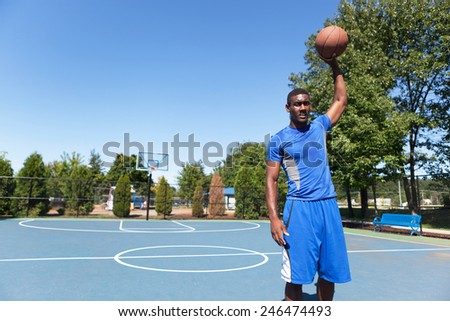 Young basketball player in his early twenties posing with the ball in one hand on an outdoor court.