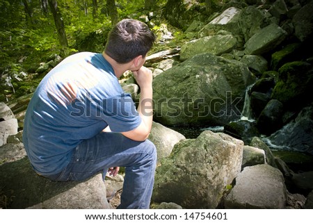 A young man in deep thought while sitting on some rocks near a beautiful waterfall.
