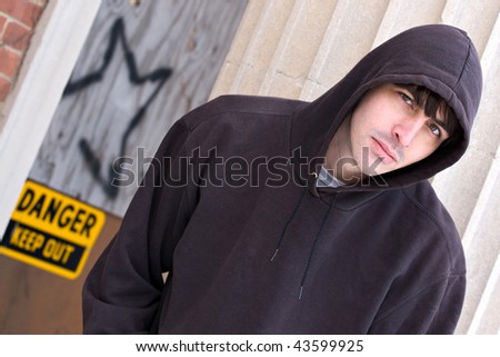 A tough looking guy posing in a grungy urban setting in a hooded sweat shirt.