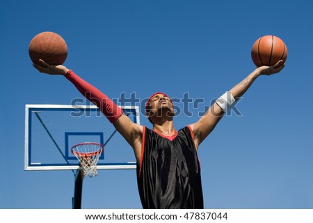 A young athletic build basketball player holding up two basketballs in the air.