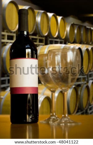 A still life shot of a single wine bottle and a pair of empty glasses in front of some stacked wine barrels.  Shallow depth of field.