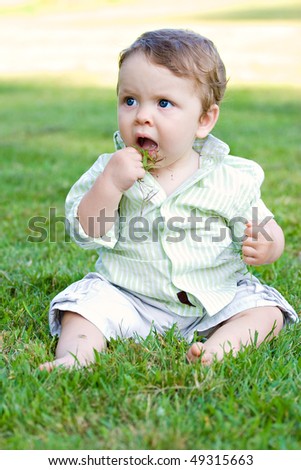 A cute baby boy about to eat a handful of green grass that he just grabbed.  He was stopped just before succeeding.