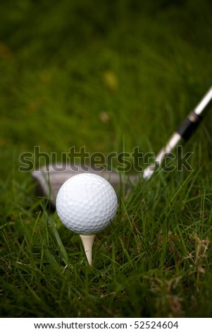 A white golf ball set up on the tee in the rough with a driver about to swing.  Shallow depth of field.