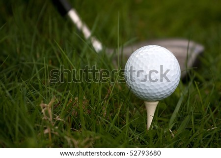 A white golf ball set up on the tee with a driver about to swing.  Shallow depth of field.