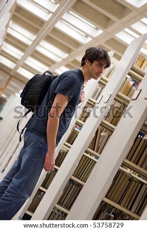 A young man reads a book at the library while standing in the aisles of book shelves.