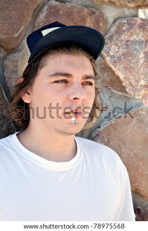 A young man smoking a cigarette wearing a white t-shirt and a trucker style hat.