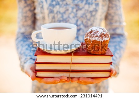 Happy woman holding pile of books, tea cup and cupcake