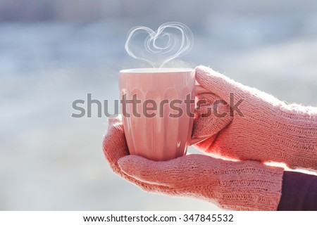 Female hands in mittens holding cup with hot tea or coffee with heart shape close up